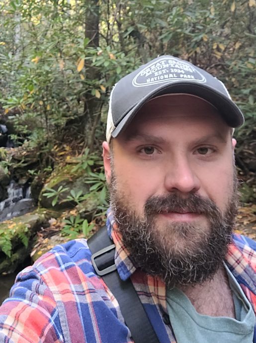 Photo of Zack Smith in the woods with a pretty waterfall behind his bearded face.